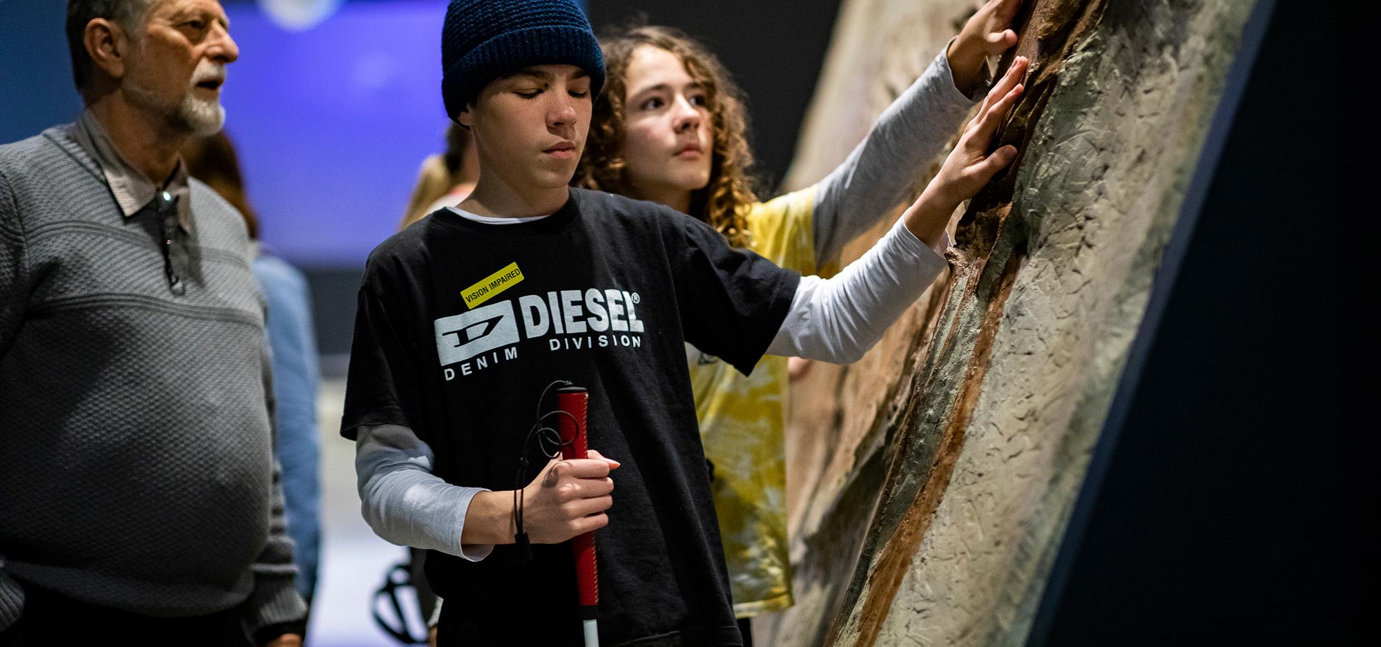 A young person wearing a blue beanie and blue t-shirt with a white Diesel logo closes their eyes as they hold a white cane in their right hand, and gently touch a textured wall with their left hand.