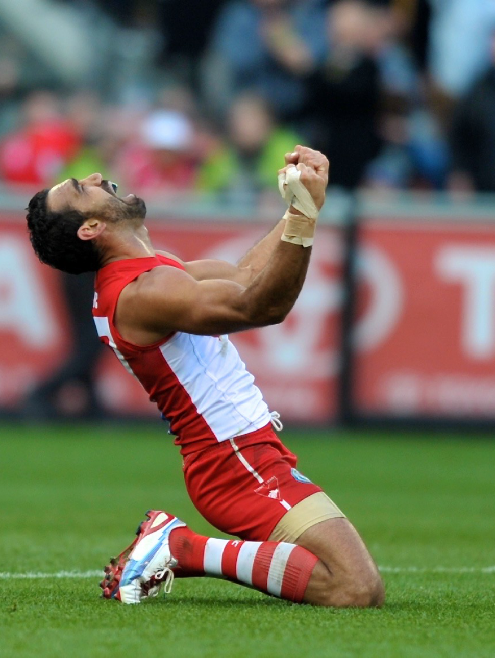 Adam Goodes is on AFL playing field, dressed in Sydney Swans uniform on his knees arms outstretched with emotion