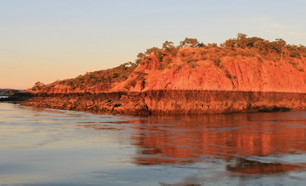 A red sunlit cliff rising up out of reflective blue water