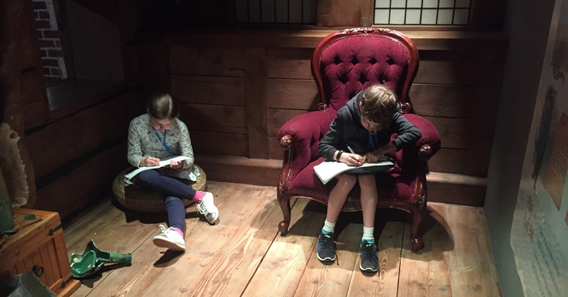 Two children sit reading, one on a wooden floor, the other on a red velvet chair