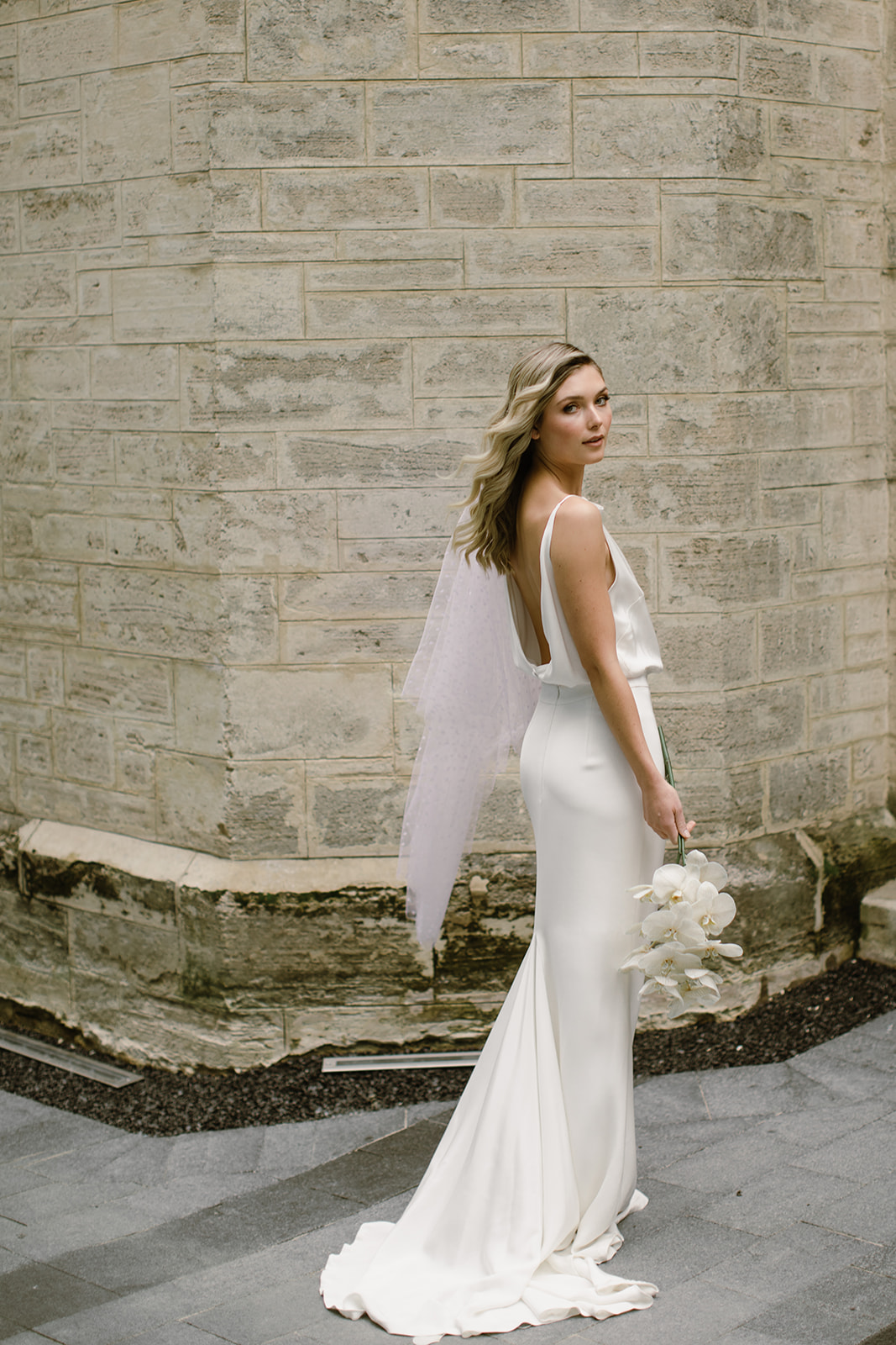 A person with blonde hair wearing a wedding dress, posing in front of a brick wall