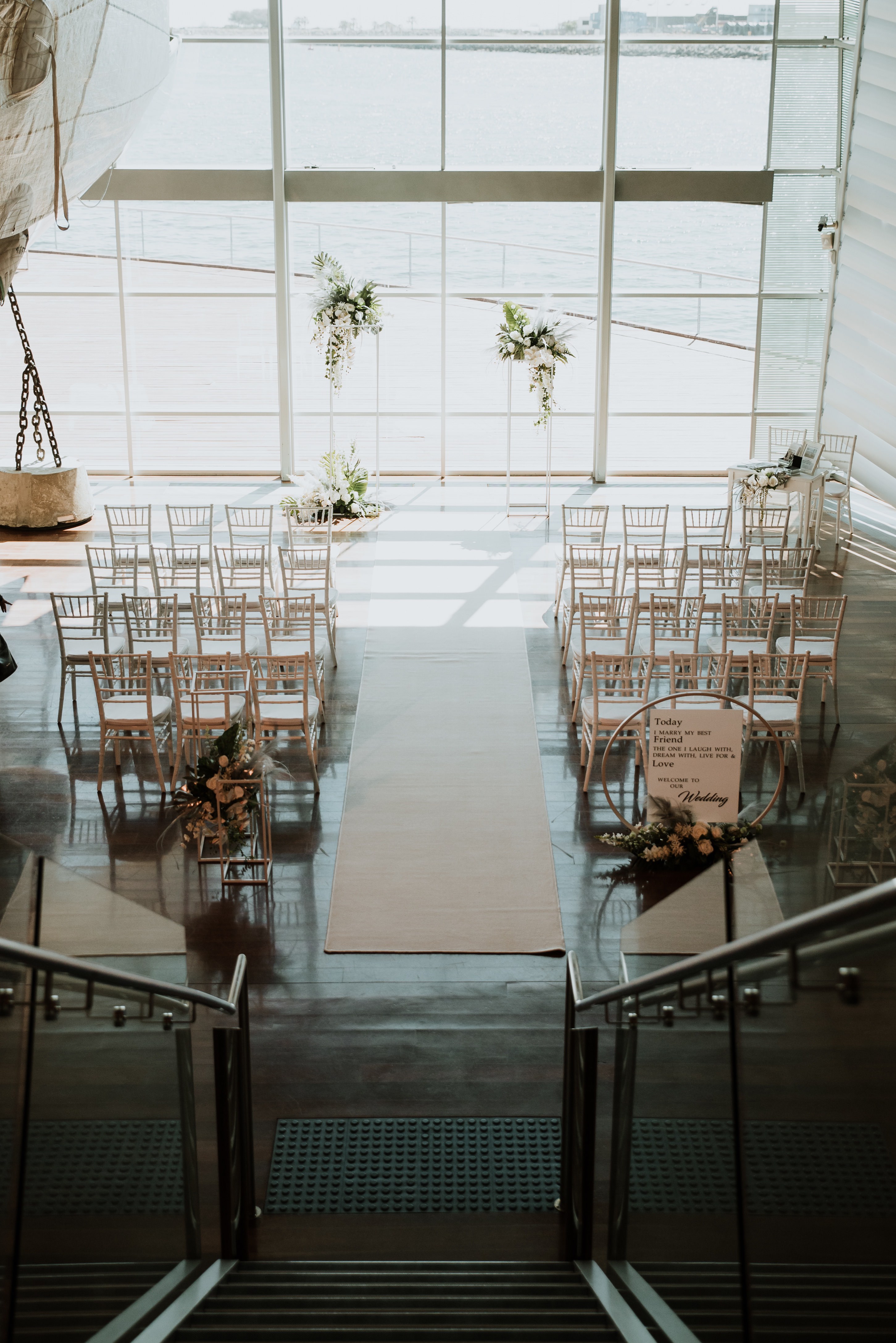 A high-angled photograph looking down a flight of narrow stairs towards two aisles of white chairs in front of a floral arch and large, brightly-lit window.