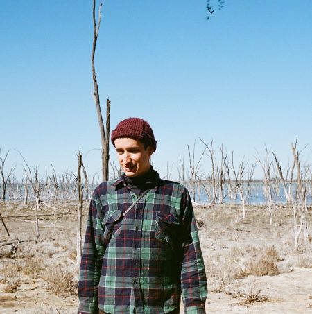 Image of a man standing in a dried lake. He wears a flannel shirt and a beanie.