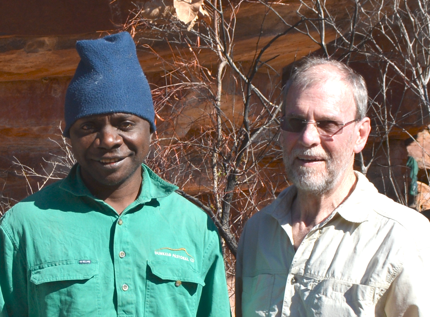 Ian Waina and Andy Gleadow from Rock Art Australia smile into the camera. Ian wears a bright green shirt and dark blue beanie and Andy wears a khaki shirt and wireframe glasses.