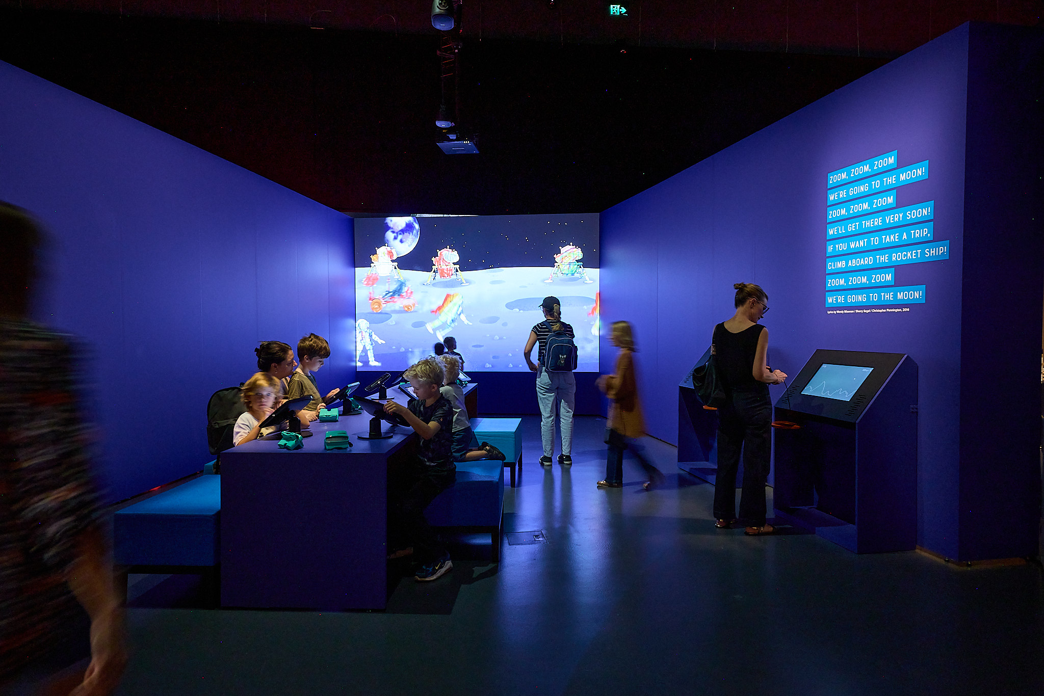 Children and adults using interactive screens in a blue-themed room with a space animation display in the background.