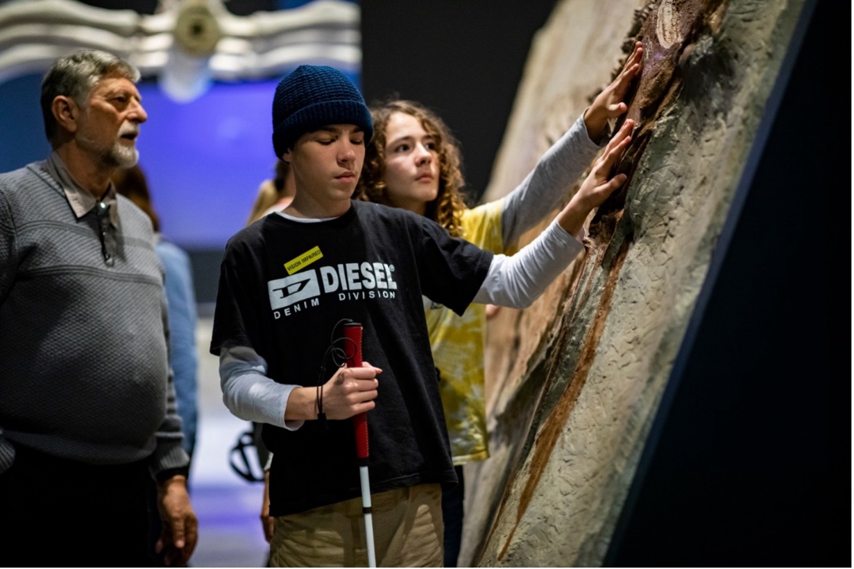 A young person with a beanie uses a white cane while touching a textured exhibit, with others nearby.