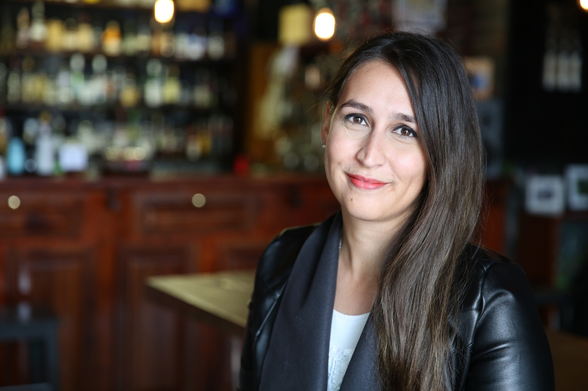 a person with long dark brown hair smiles at the camera wearing a white top and black jacket