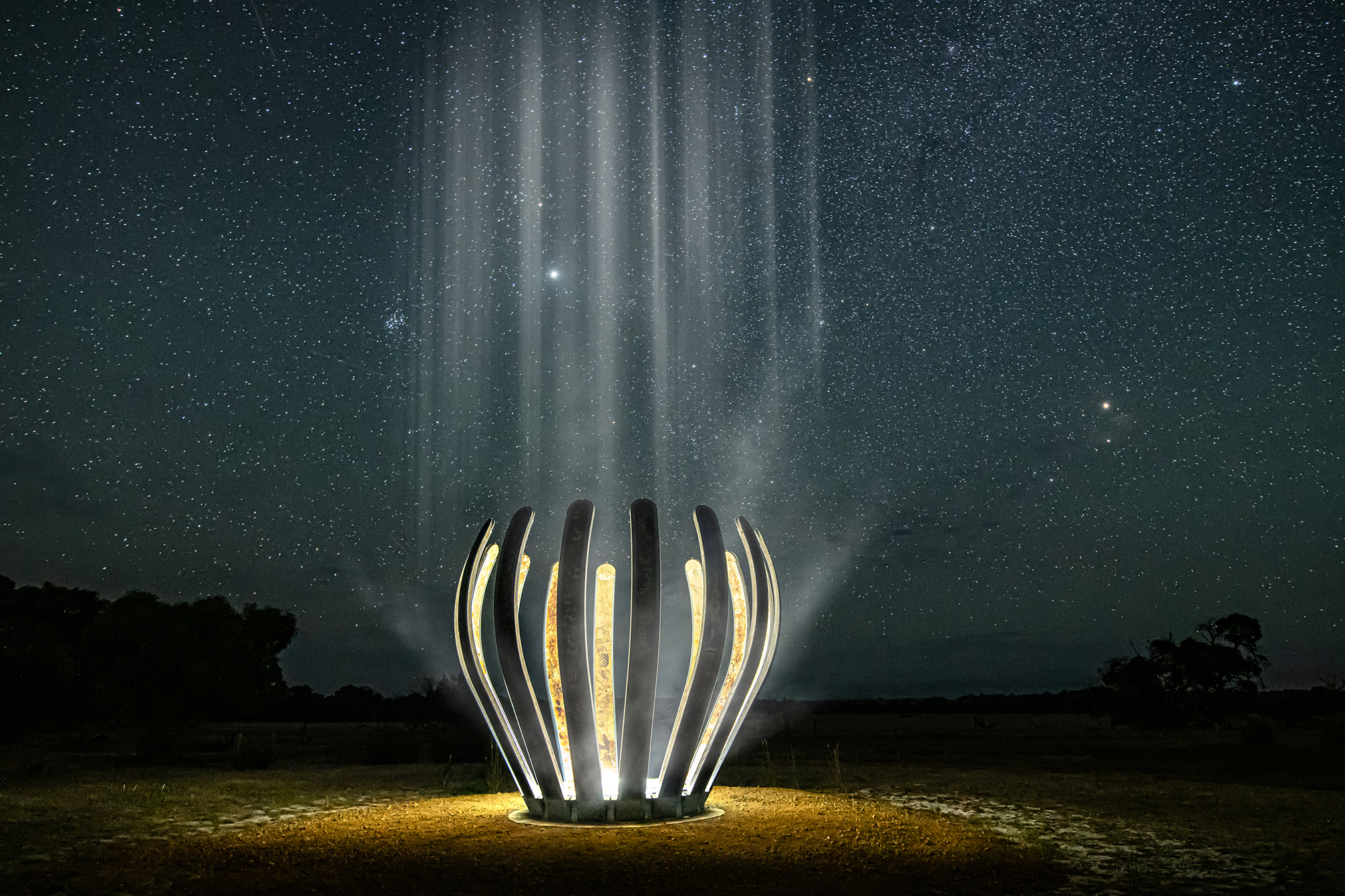 Illuminated sculptural artwork glowing on open land at night, with beams of light rising into a star-filled sky.