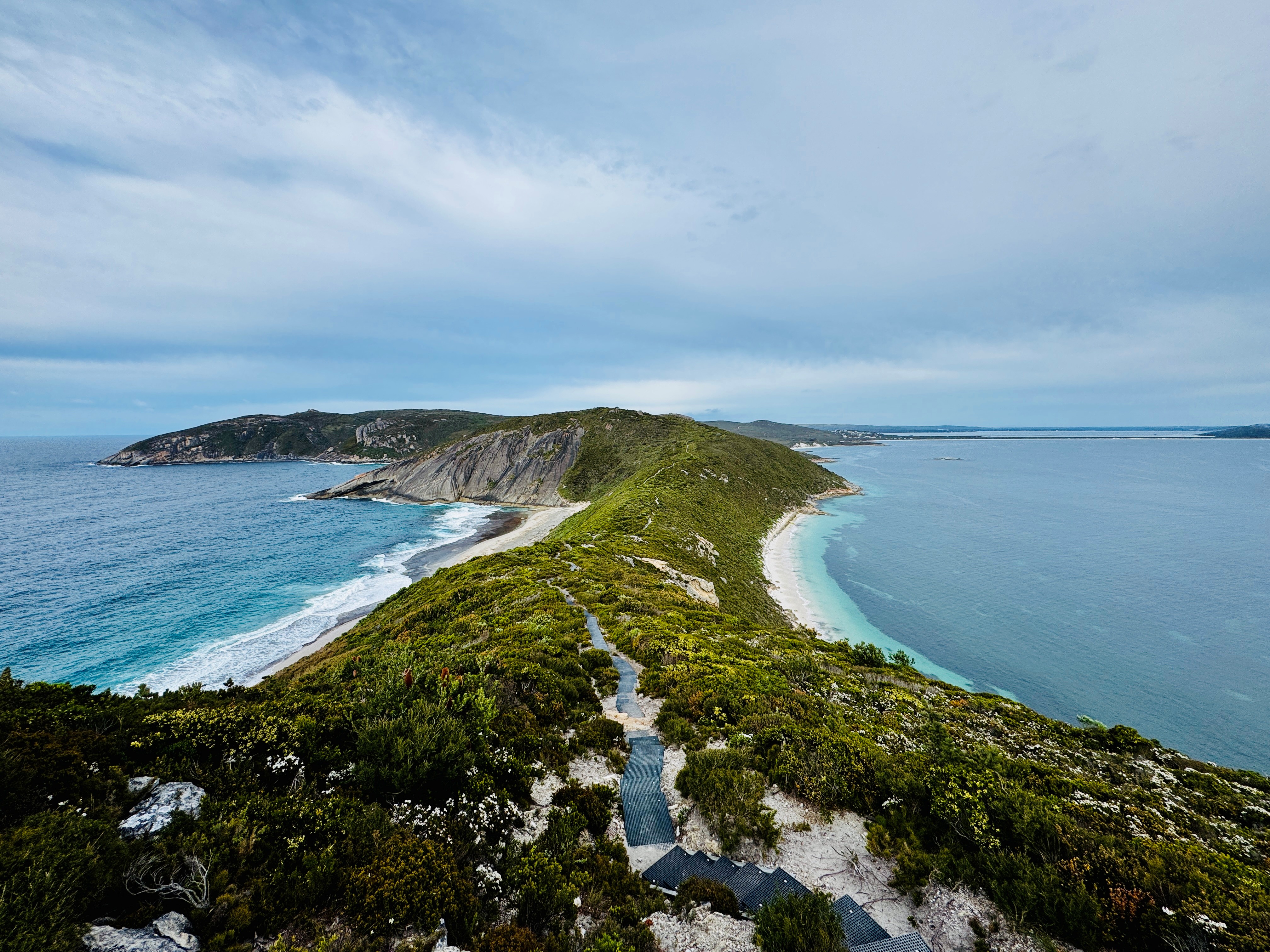 Elevated view of a narrow coastal headland with a walking path, flanked by blue ocean on one side and turquoise water on the other under a wide sky.