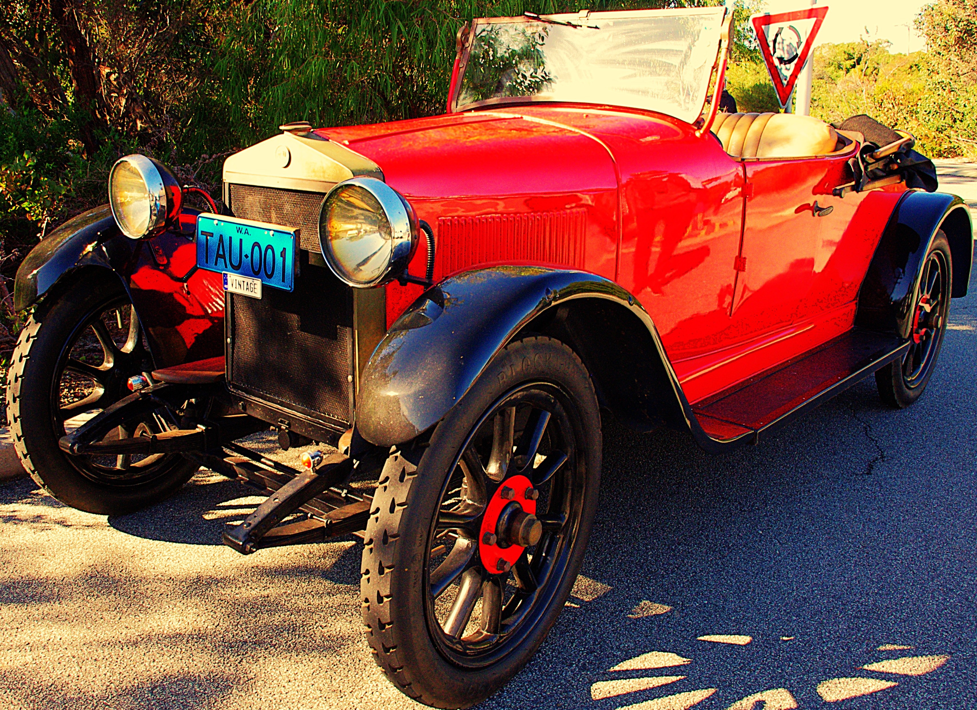 Vintage red open-top car parked on a roadside, with black wheels, round headlights, and a visible blue license plate in warm sunlight.