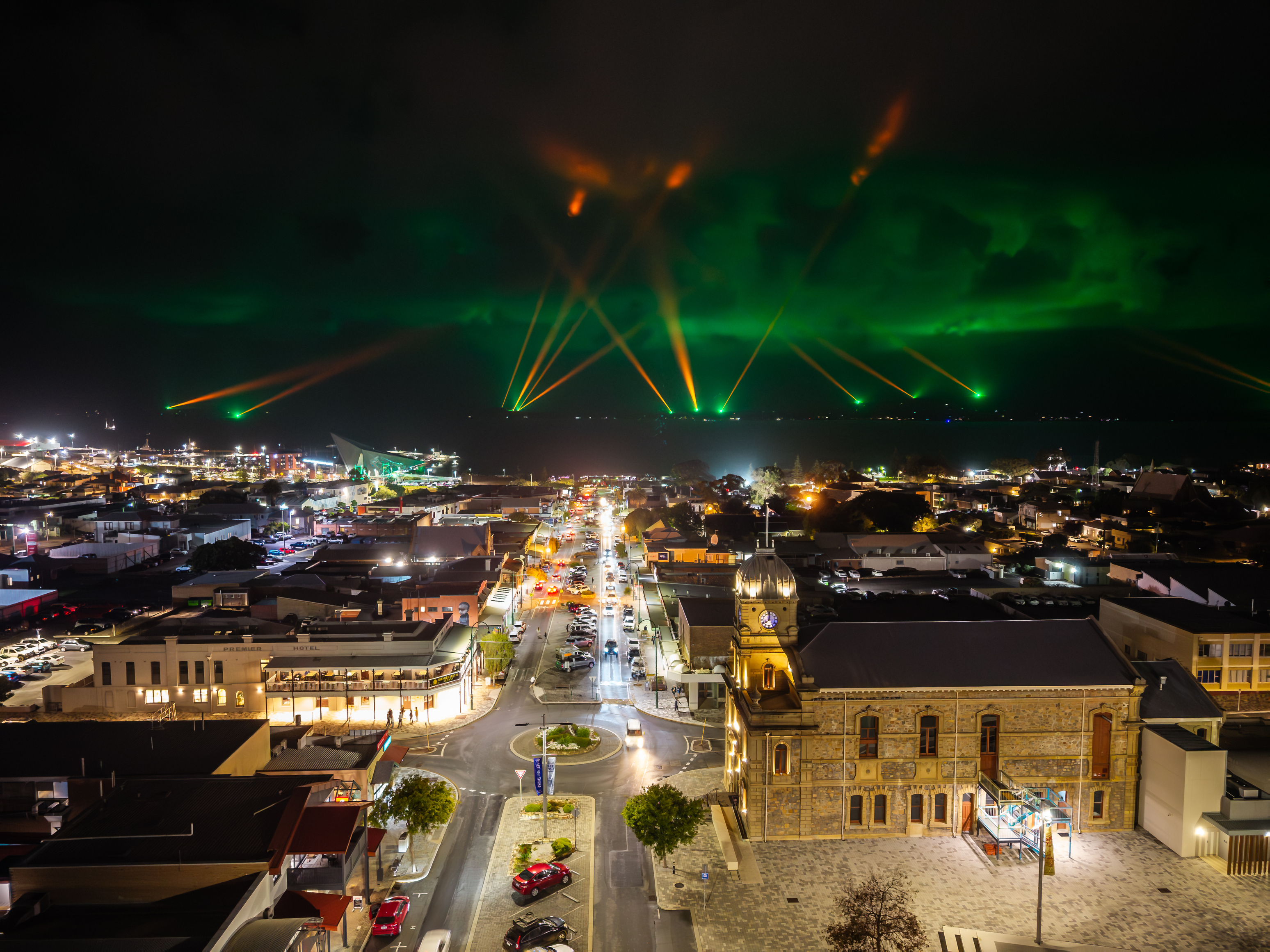 Nighttime aerial view of a coastal town with lit streets and historic buildings, while green and orange laser lights beam across the sky over the water.