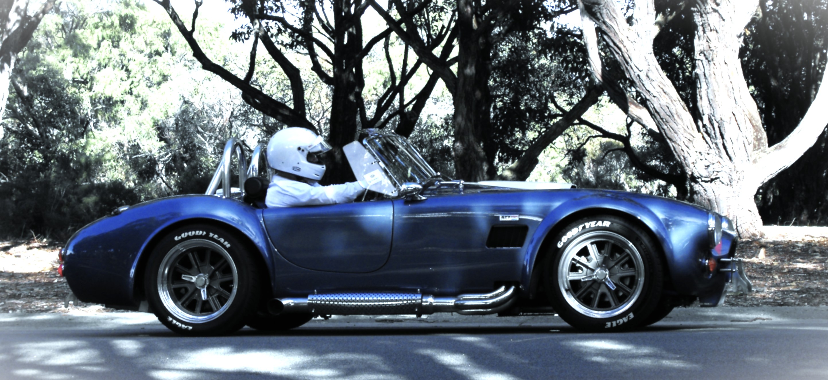 Side view of a blue open-top sports car on a tree-lined road, with a helmeted driver seated behind the wheel.