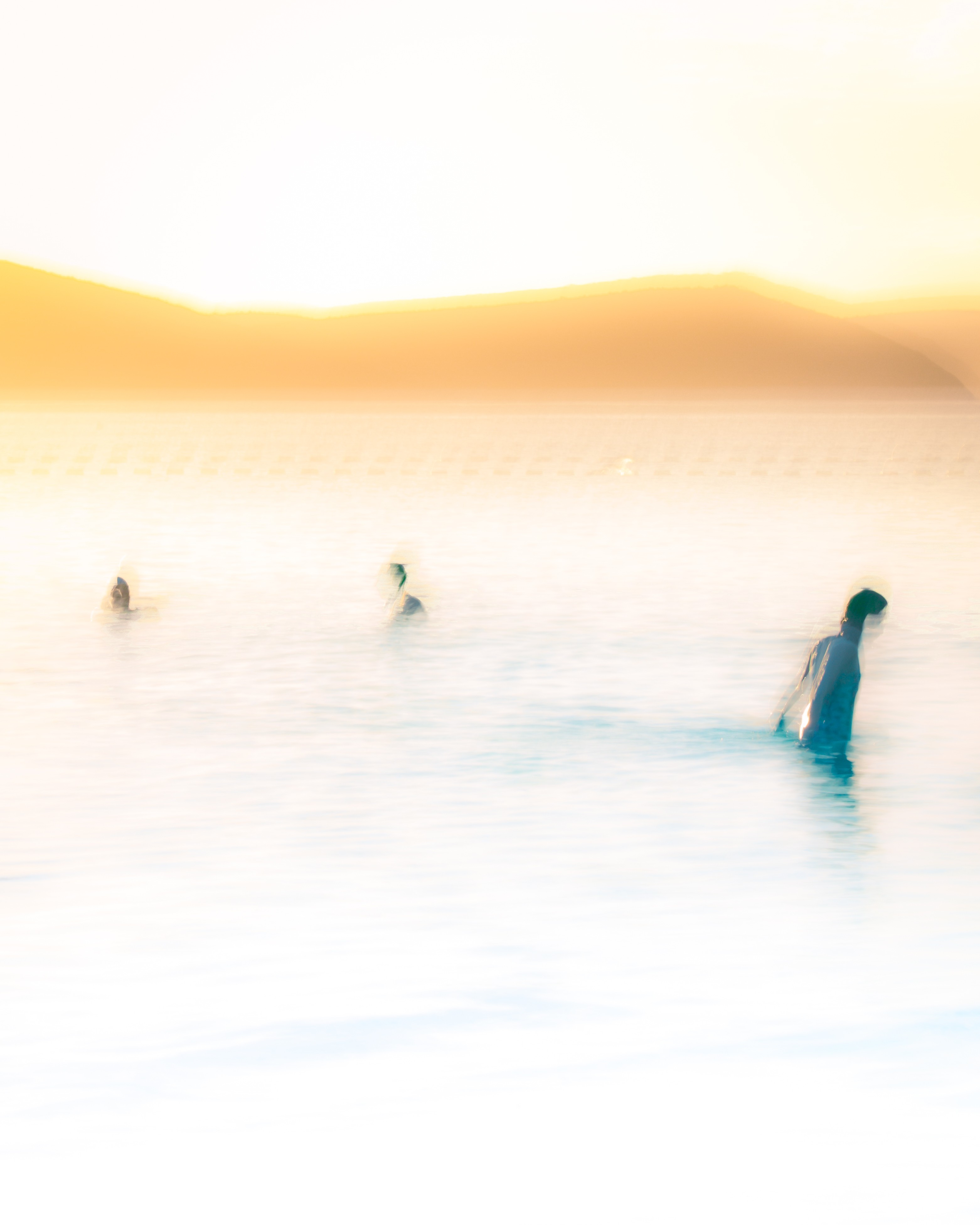 Soft, hazy scene of people wading in calm water at sunrise, with warm golden light and distant hills in the background.