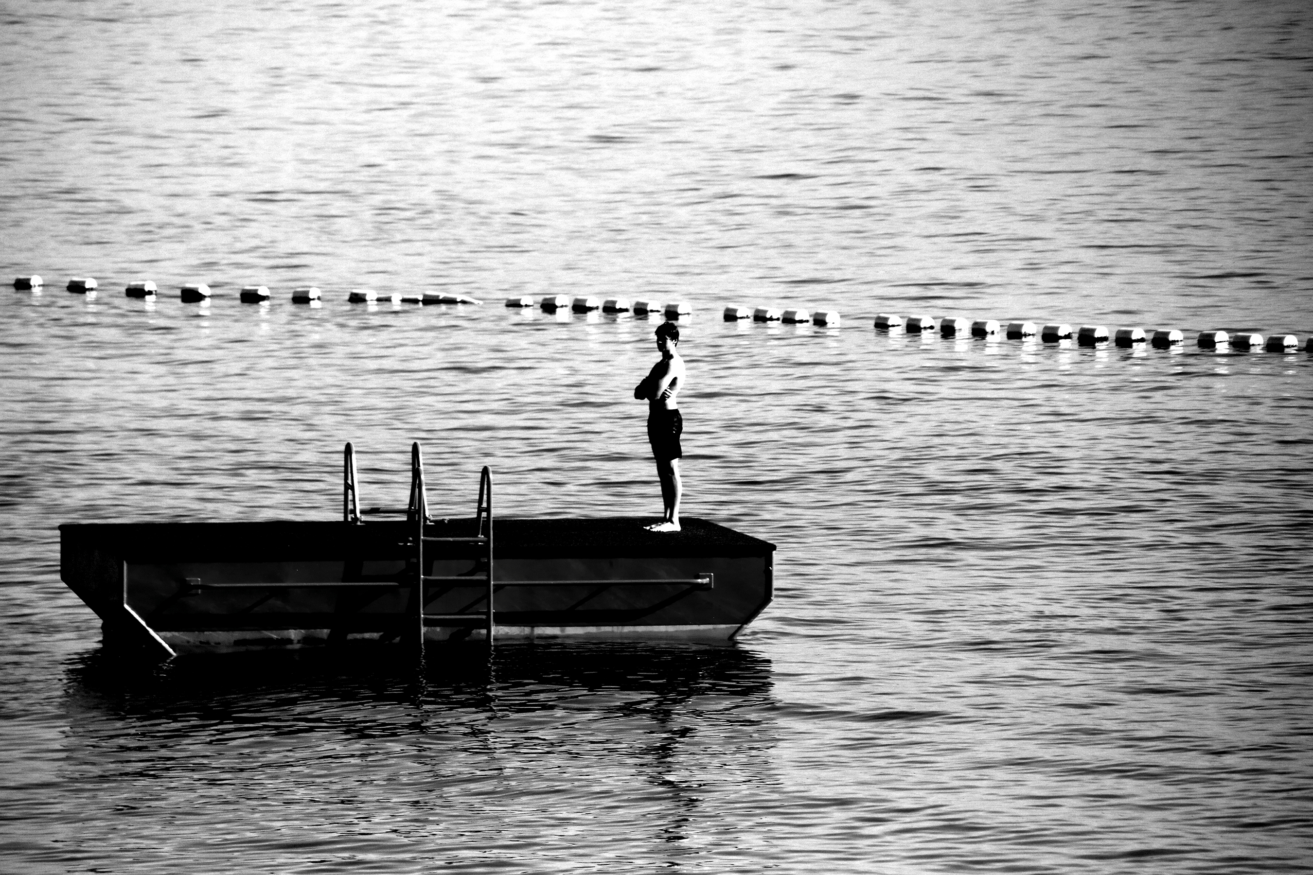 Black-and-white image of a person standing on a floating pontoon with a ladder, surrounded by calm water and a line of buoys in the distance.