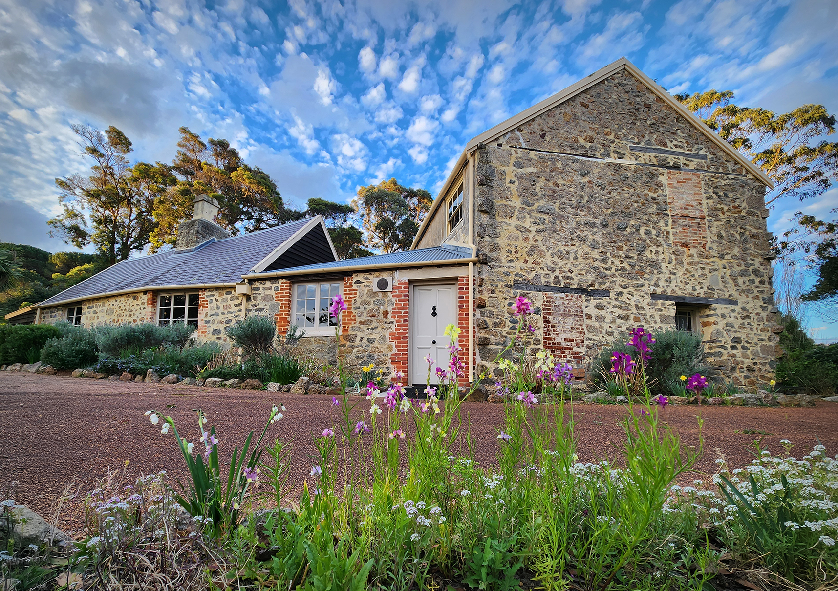Stone farmhouse with red brick details and a pitched roof, surrounded by garden flowers under a bright blue, cloud-dotted sky.