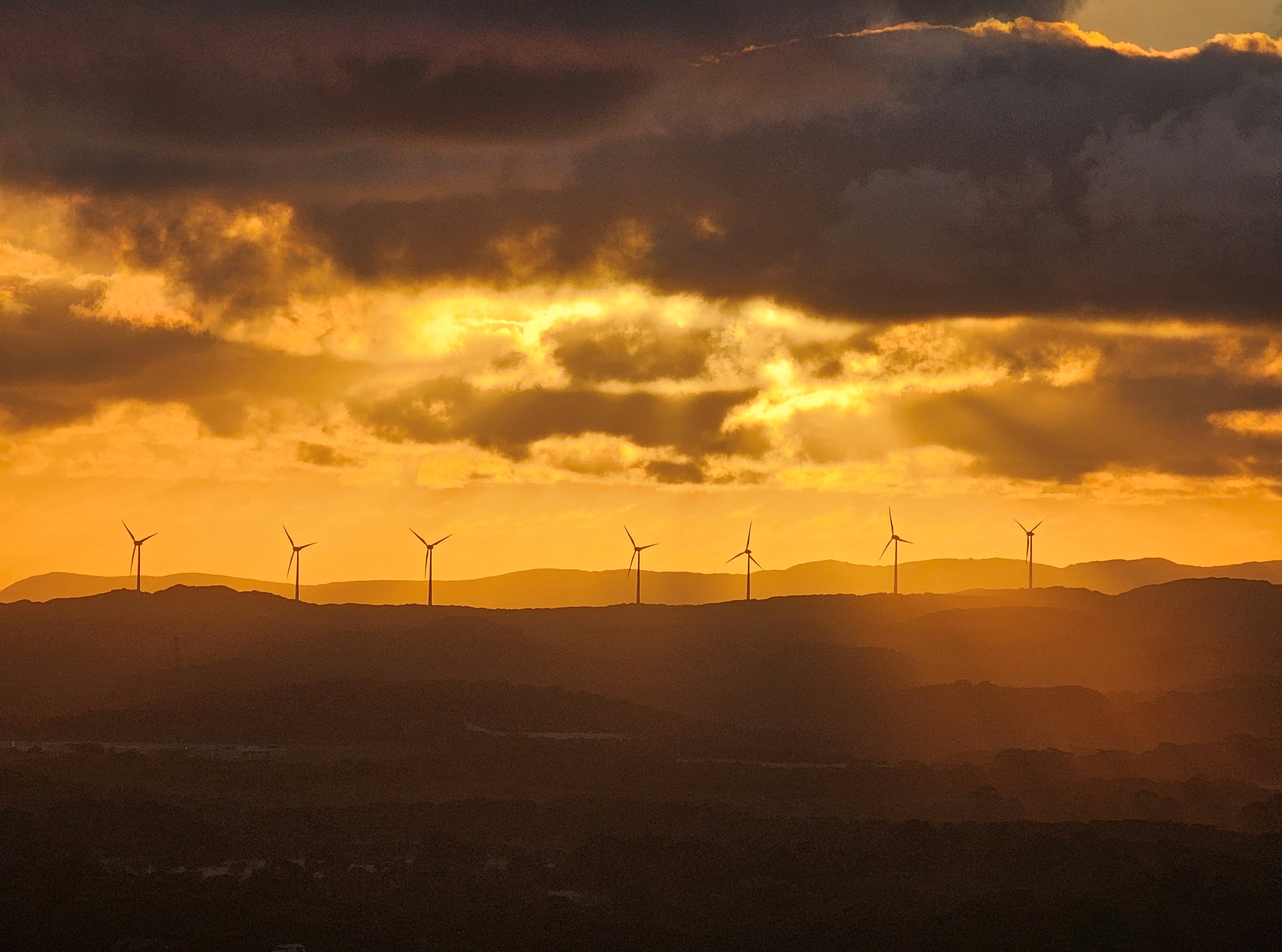 Silhouetted wind turbines on a ridgeline against a vibrant golden sunset with dramatic clouds.