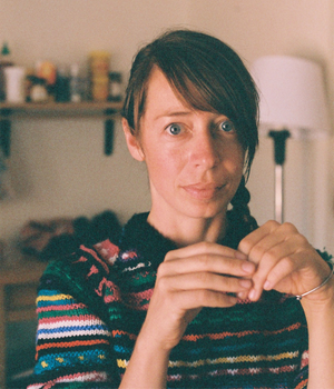 A person wearing a colourful striped knitted sweater sits indoors, looking toward the camera with hands raised near chest level. Shelves and a floor lamp are softly blurred in the background.