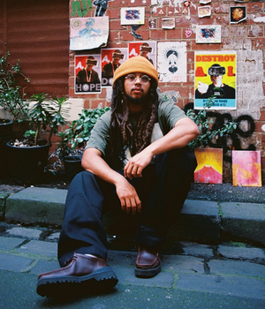 Man with long hair and beanie sitting down in front of art/music posters