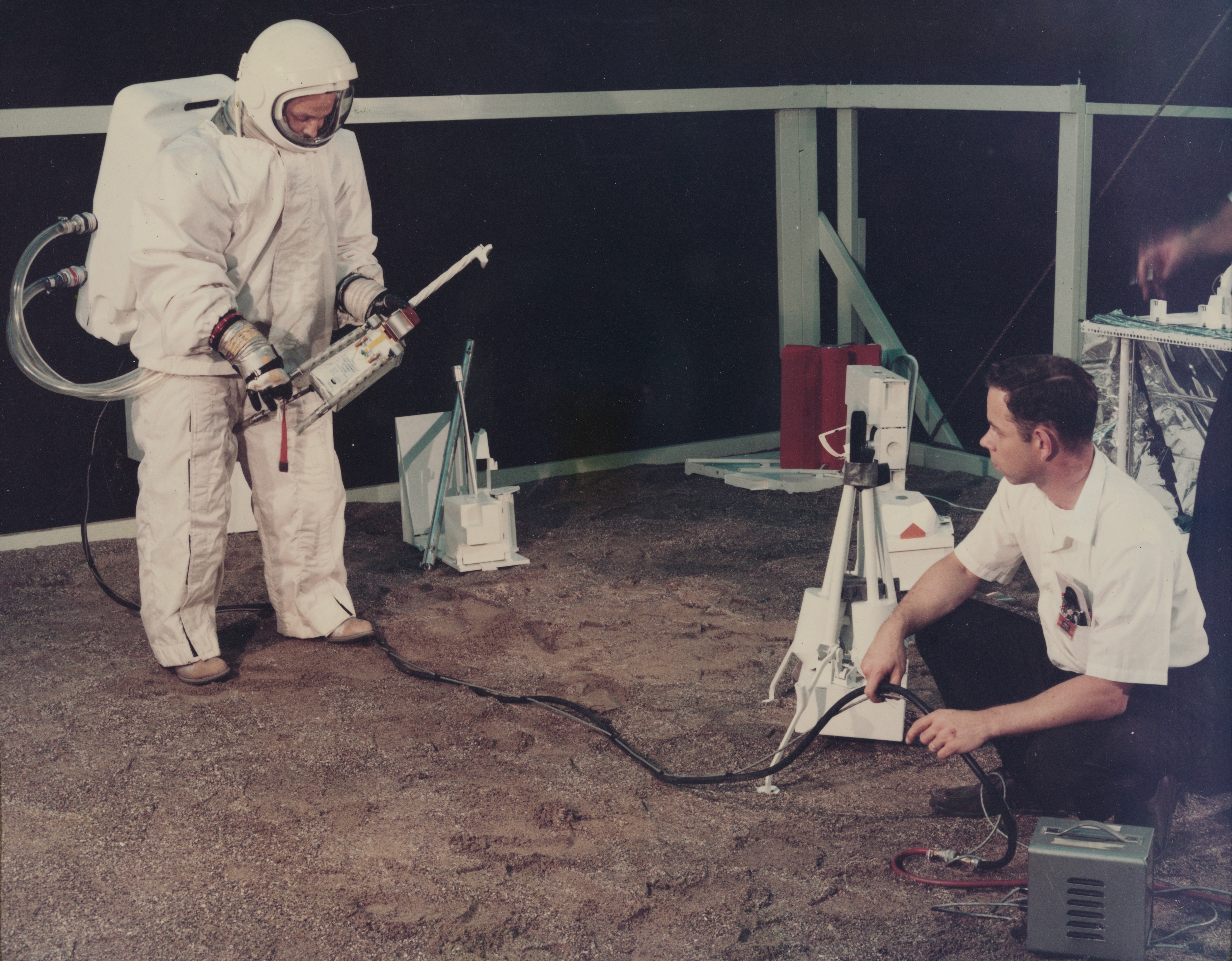 An old image of a man in a large bulky white astronaut suit holding a large instrument with a long spout while another man crouches on the floor, watching