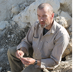 A person sits outside on rocks dressed in light coloured clothing looks directly at camera