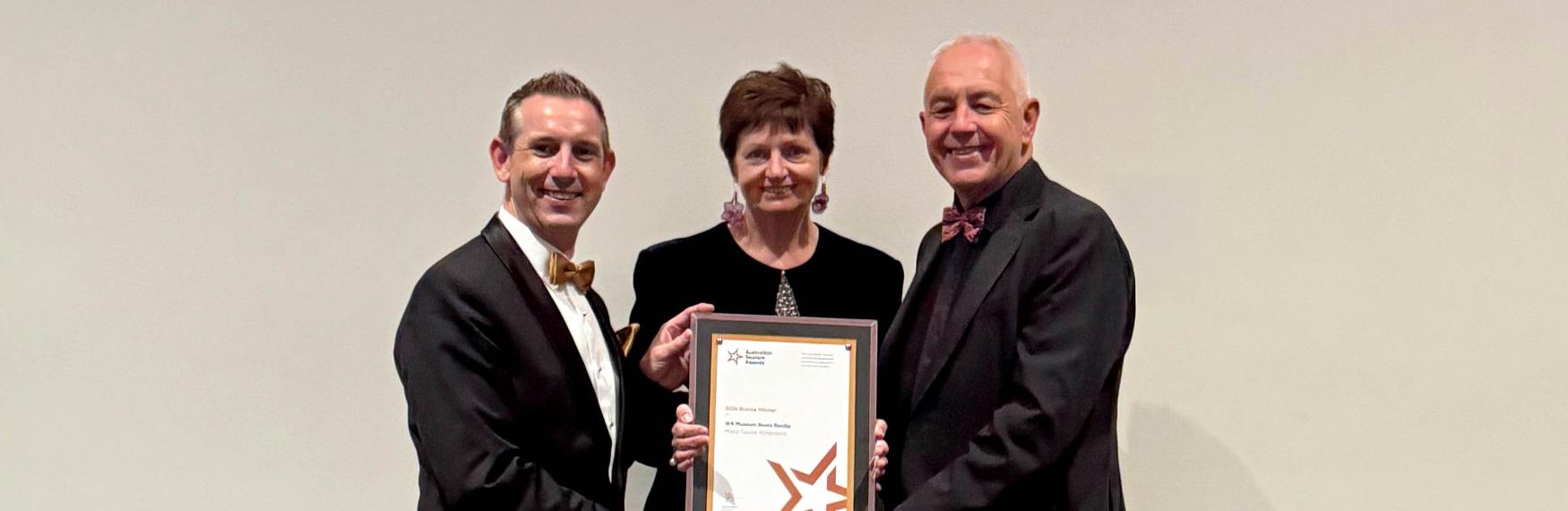 Two men and a woman, all wearing black formal wear, hold a Bronze Winning certificate.