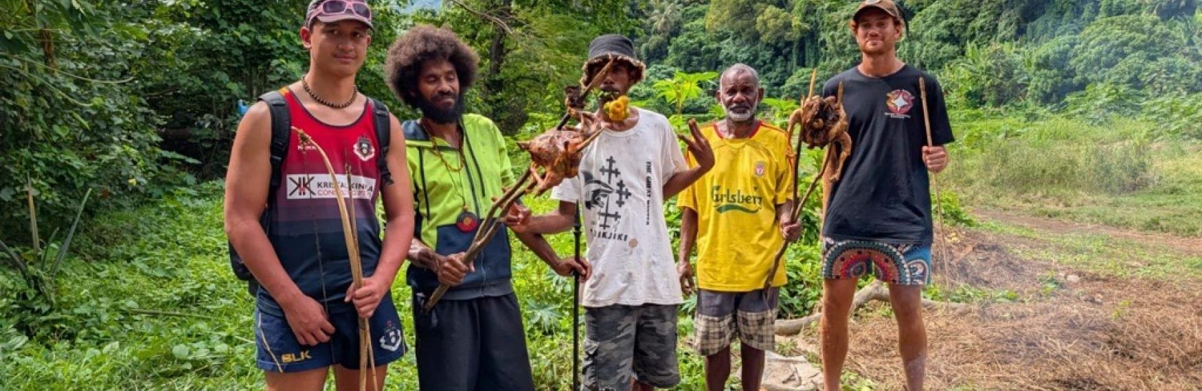 Five Indigenous young people standing around a small fire holding sticks with meat while surrounded by greenery