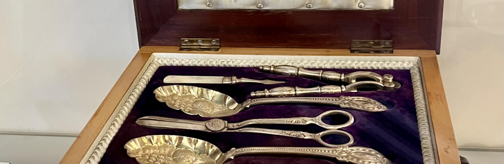 An open wooden presentation box with a satin-padded lid and a deep purple velvet interior, displaying an ornate Victorian-era silver serving utensil set. The set includes intricately designed spoons, tongs, and scissors with decorative handles, arranged neatly inside the box.