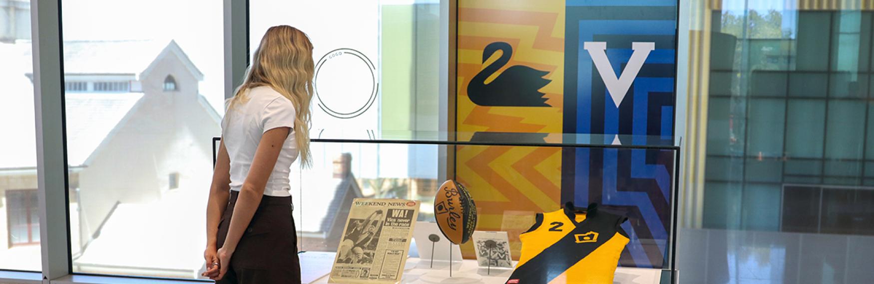 A woman looks at a small showcase of AFL memorabilia in a Museum. The showcase is surrounded by the WA and Victorian Football logos.