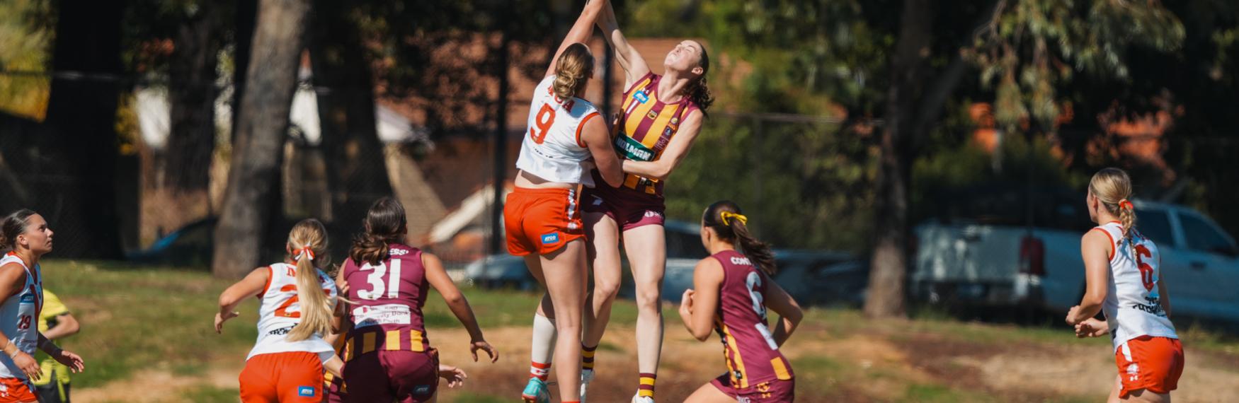 A group of female AFL players mid game, with two jumping high for a ball.
