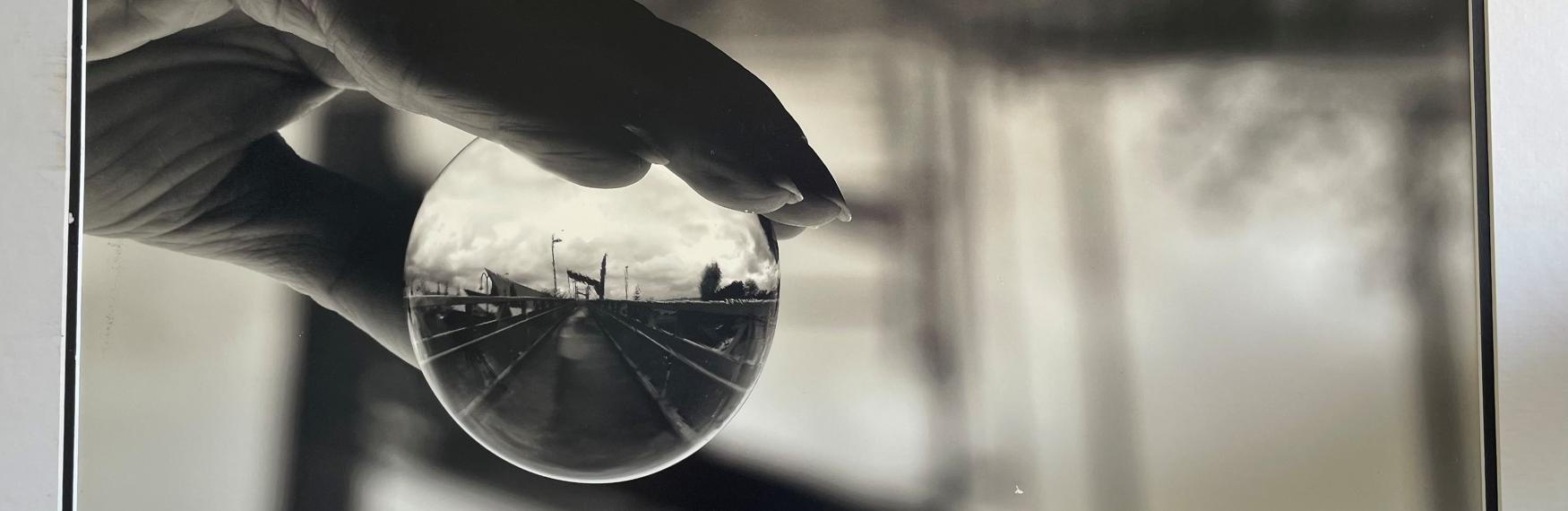 Black-and-white photo of a hand holding a glass sphere that reflects an upside-down view of a pier and cloudy sky.