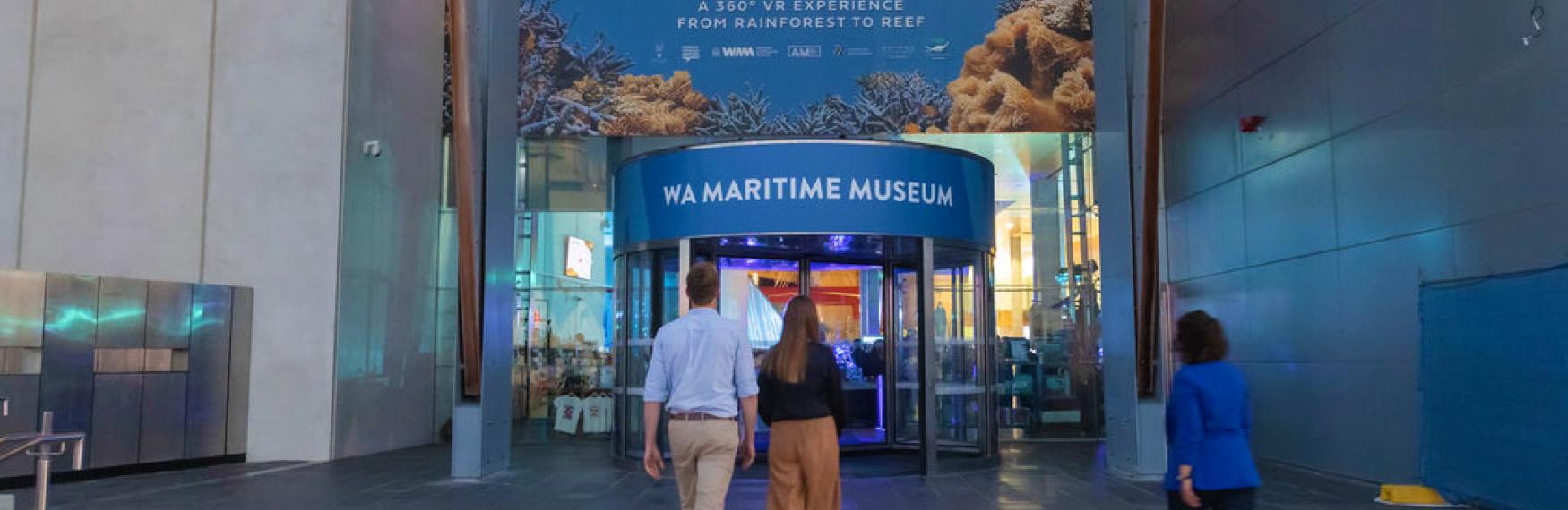 Three people walking to the entrance of the WA Maritime Museum, displaying advertising for Life in the Great Barrier Reef