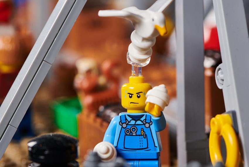 A LEGO seagull perches on the head of a LEGO man who is trying to eat an ice cream