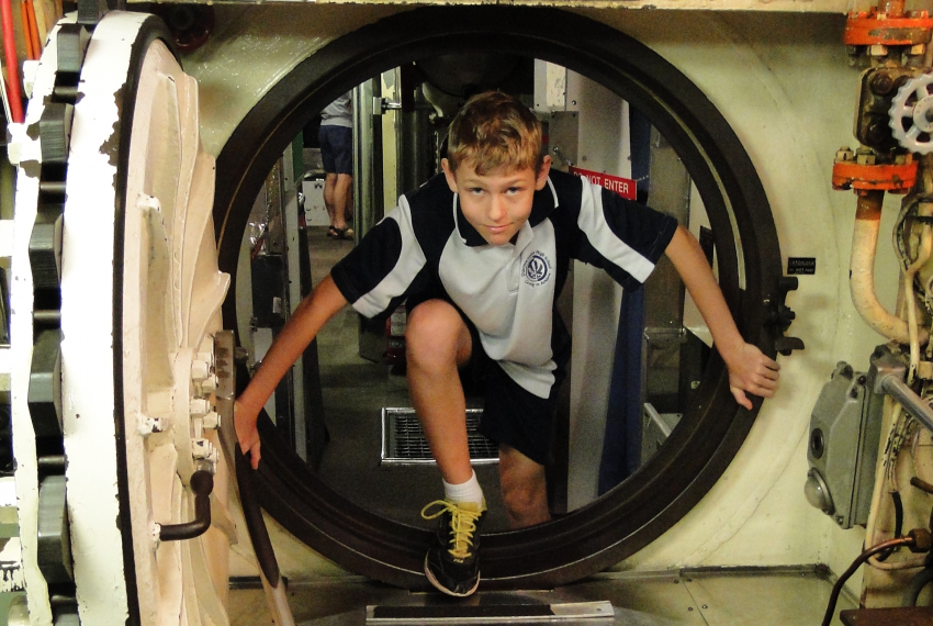 A boy dressed in school uniform climbs through a small hatch door in a submarine