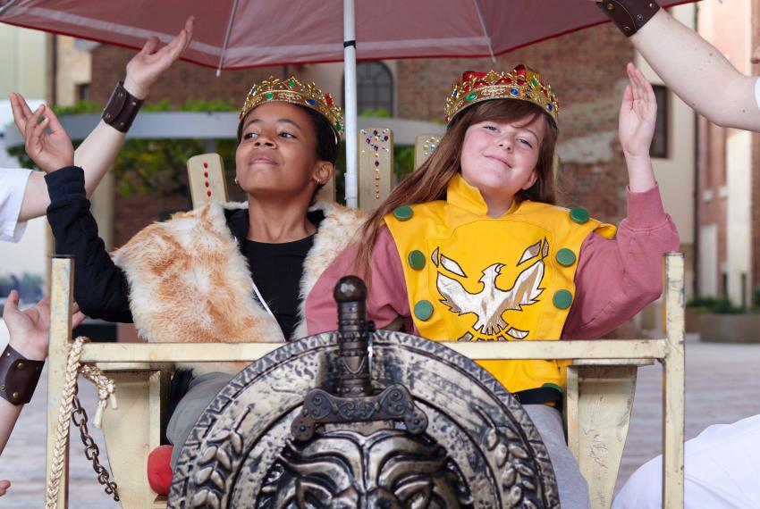 Two children dressed up like royalty sit in a chariot and wave grandly to the onlookers