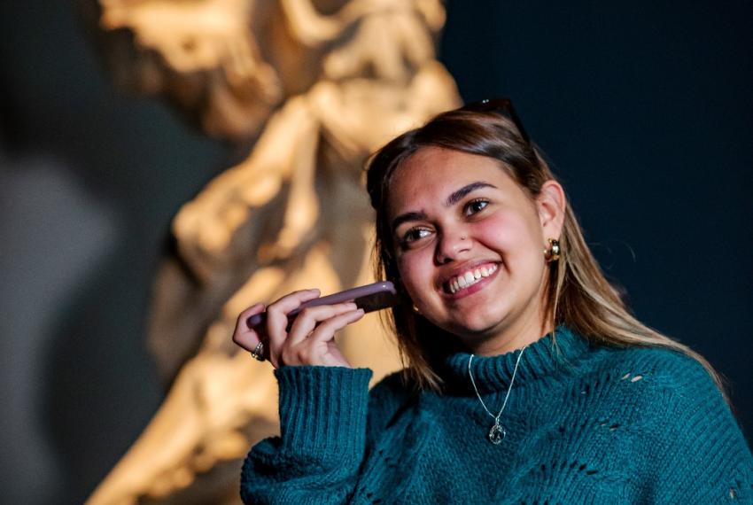 A woman standing in front of an Ancient Greek statue holds a smartphone up to her ear as though listening