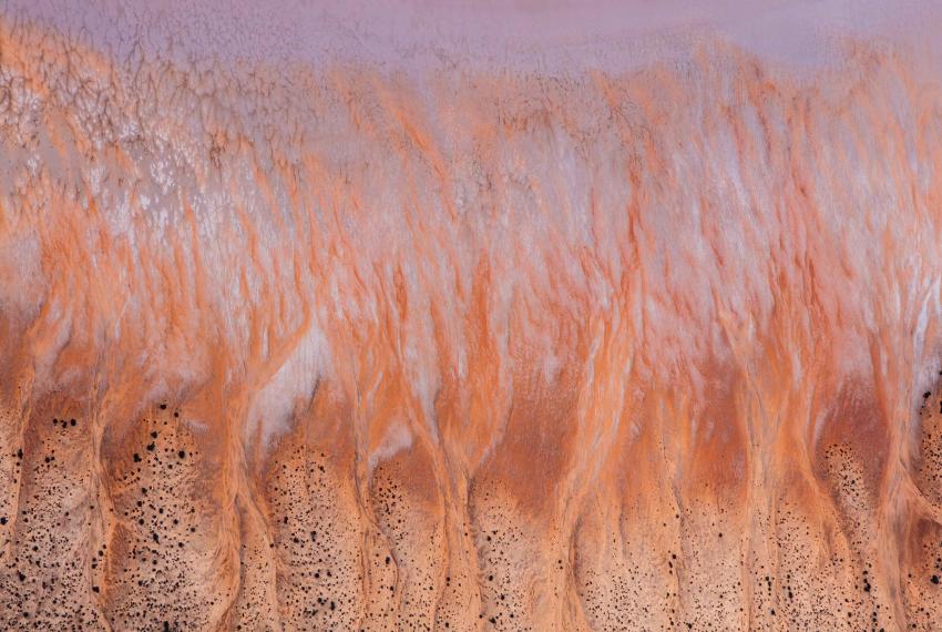 An aerial view of a hilly striated salt lake landscape in shades of red, yellow and white