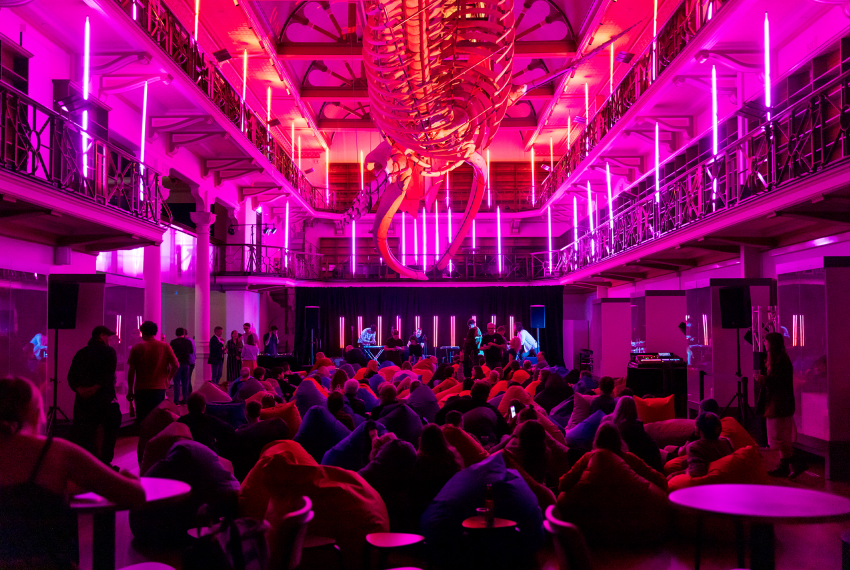 An audience seated below a suspended whale skeleton watch a music performance in a dramatically-lit heritage building