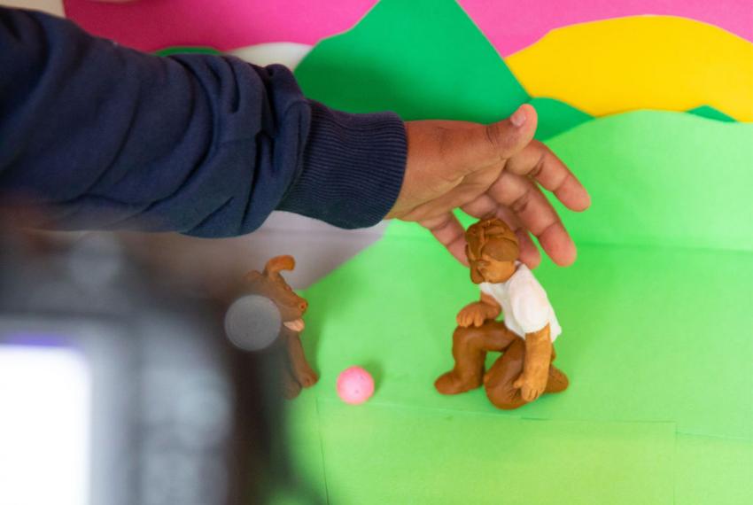 A child's hand reaches out to adjust a clay figure against a bright green backdrop