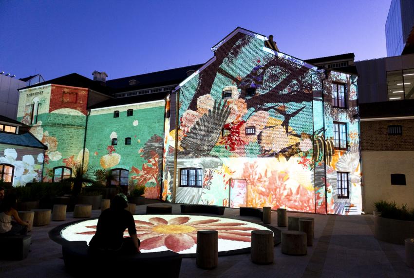 Night time shot of the Old Goal Building, on which the Illuminations projection is being played. At the forefront of the image is the Dance Circle, which is also lit up by projected artwork. 