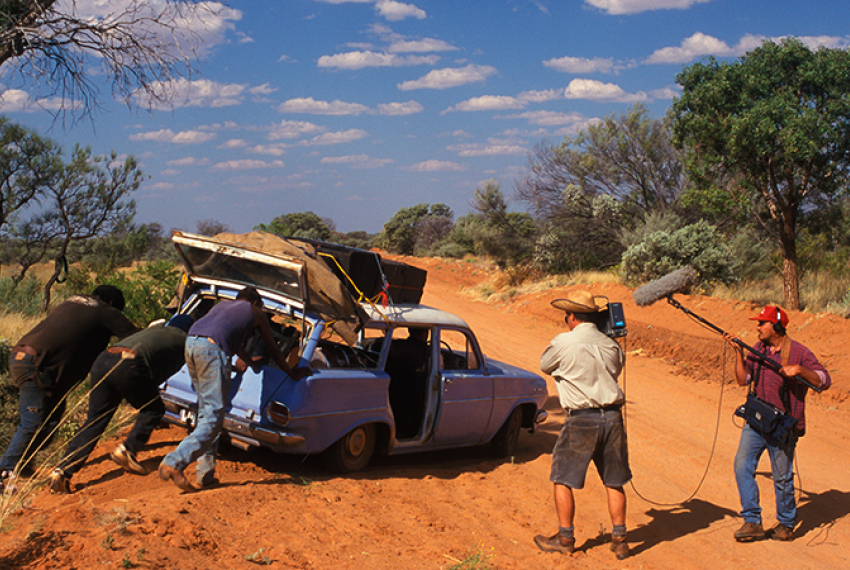 Three people push on an old, blue car bogged on a dirt track, whilst 2 camera people film.