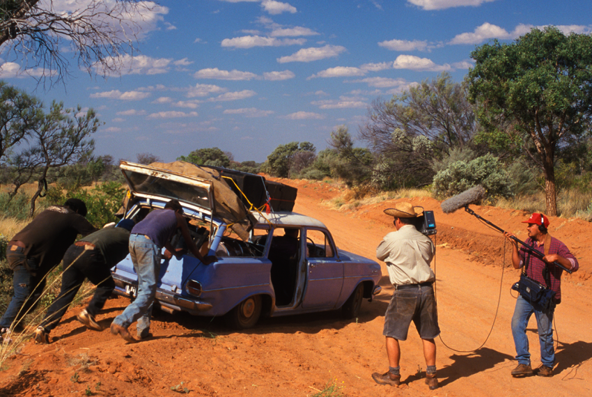 3 people push an old, blue car bogged on the side of a bush track, while 2 camera people film.