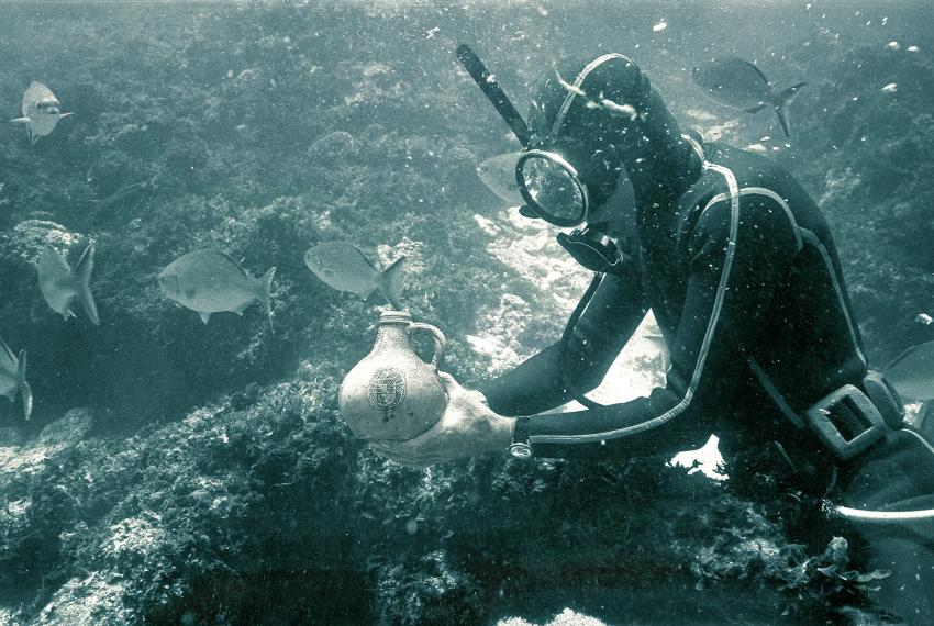 A diver underwater at a shipwreck site examines a pottery jug