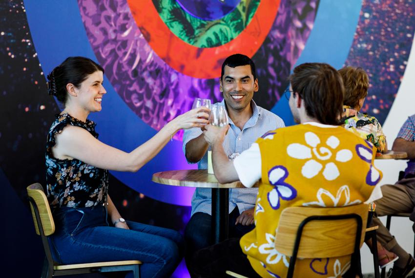 Three people are sitting at a high top table. One has their back to us. They have smiles on their faces and are in the middle of a cheers gesture with their drinks 