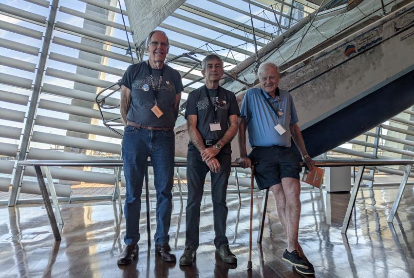 Three mature male volunteers wearing WA Museum tee shirts and lanyards stand in front of a sailing vessel on display in the WA Maritime Museum, smiling at the camera