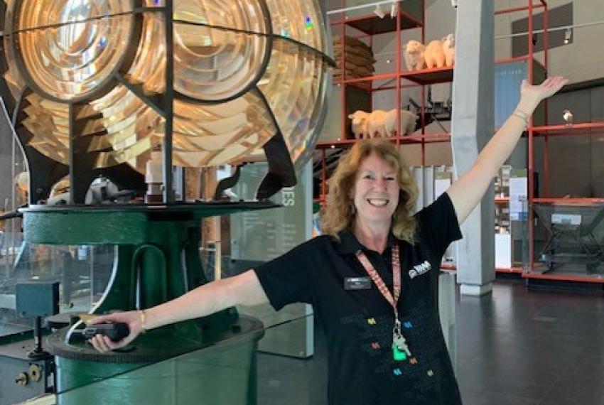 A female volunteer wearing a WA Museum tee shirt smiles and poses with her arms outstretched in front of a large lighthouse lamp