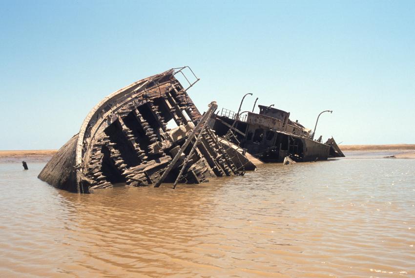 A shipwreck in shallow water near a sandbar, beneath a light blue sky. Most of the upper half of the shipwreck is exposed to the air above the water's surface.