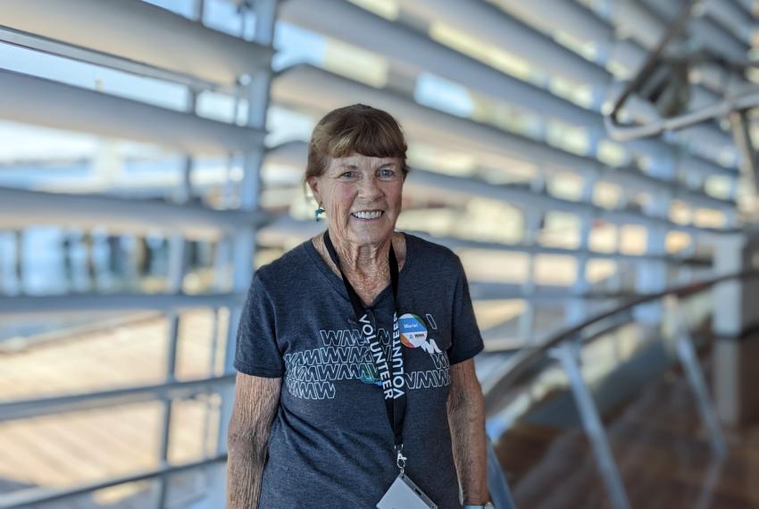 A female volunteer wearing a WA Museum tee shirt stands in the WA Maritime Museum, smiling at the camera