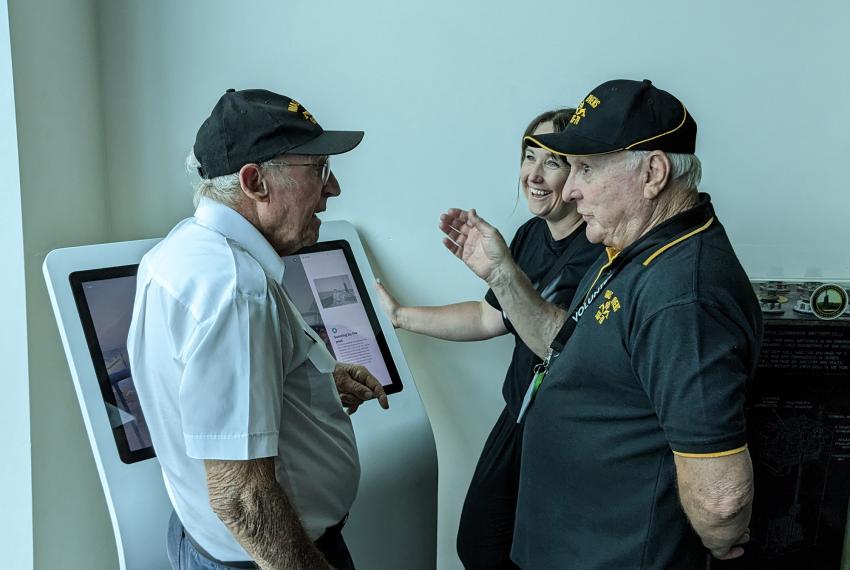 Three people stand near a digital display in a museum, talking together and laughing