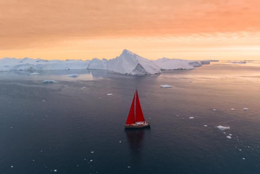 boat with red sails is sailing in water with icebergs in the background