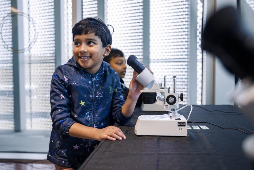 A child with black hair and a rain jacket with colourful stars is seated at a microscope, turning to their right, towards the camera, to smile at something over their shoulder